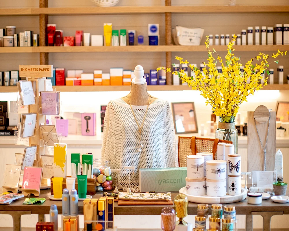 A cozy boutique with a mannequin centerpiece, shelves of colorful skincare and gifts, bright lighting, and a vase of yellow blossoms on a wooden display.