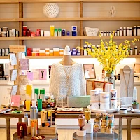 A cozy boutique with a mannequin centerpiece, shelves of colorful skincare and gifts, bright lighting, and a vase of yellow blossoms on a wooden display.