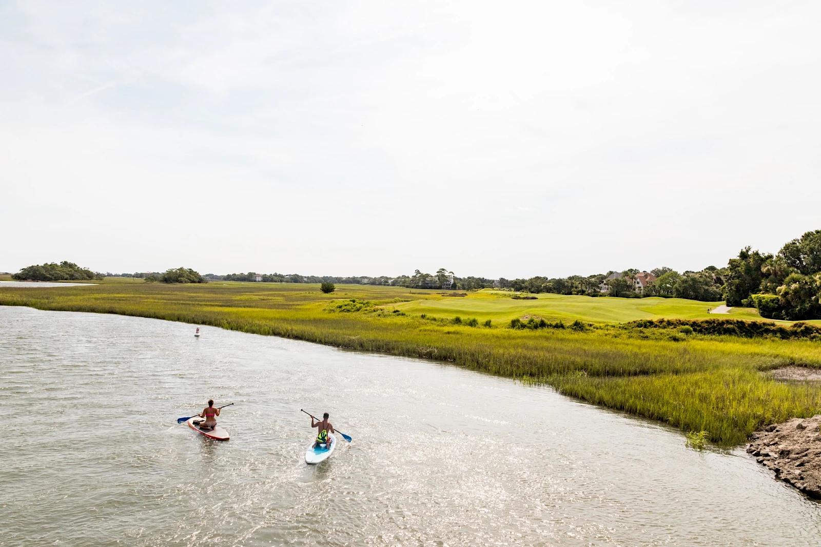 Two people paddleboarding in a calm river, grassy marshland on the bank under a bright sky.