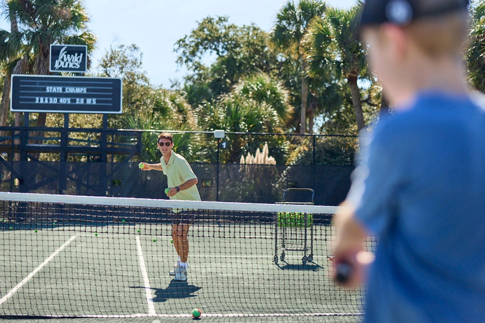 Two people playing tennis on a sunny court, with one about to serve and a scoreboard in the background.