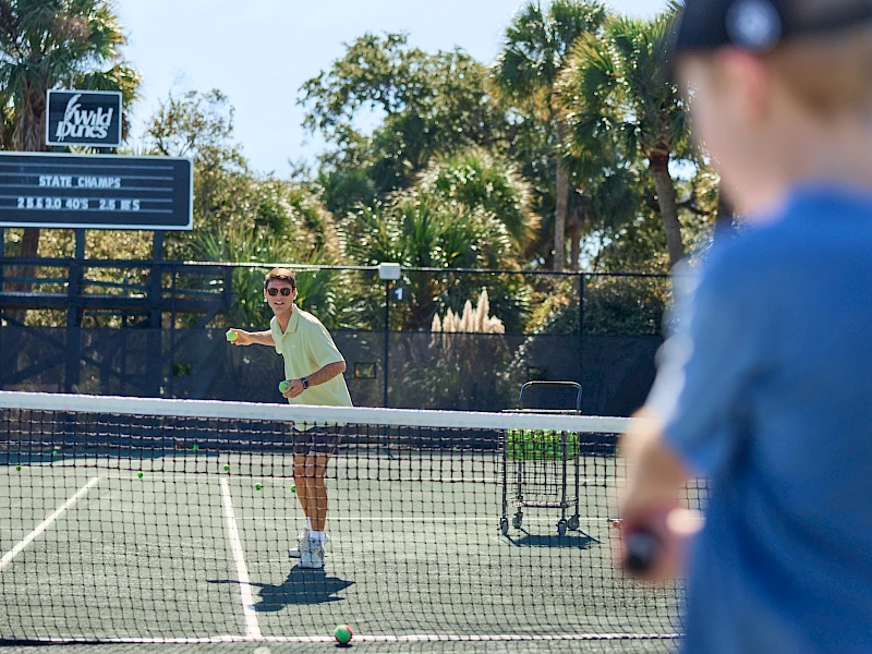 Two people playing tennis on a sunny court, with one about to serve and a scoreboard in the background.