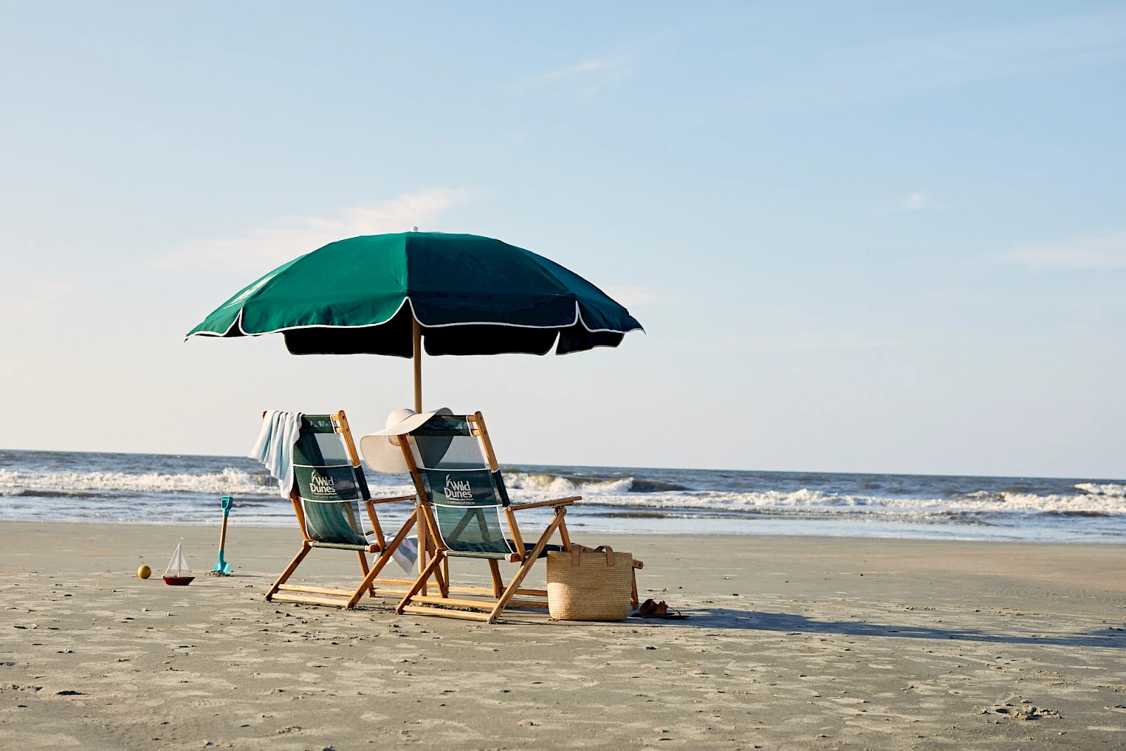 Two empty beach chairs under a green umbrella on calm sand by the sea, with a bucket and shovel nearby.