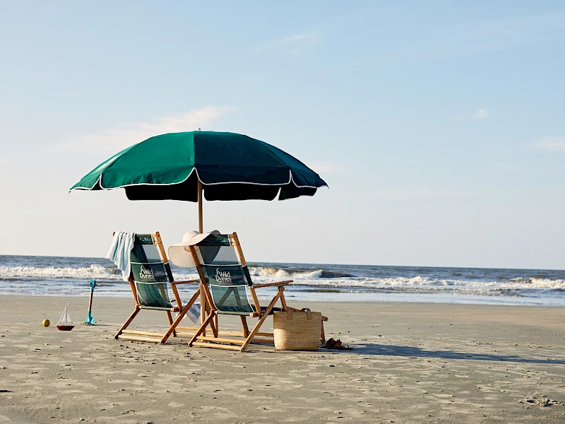 Two empty beach chairs under a green umbrella on calm sand by the sea, with a bucket and shovel nearby.
