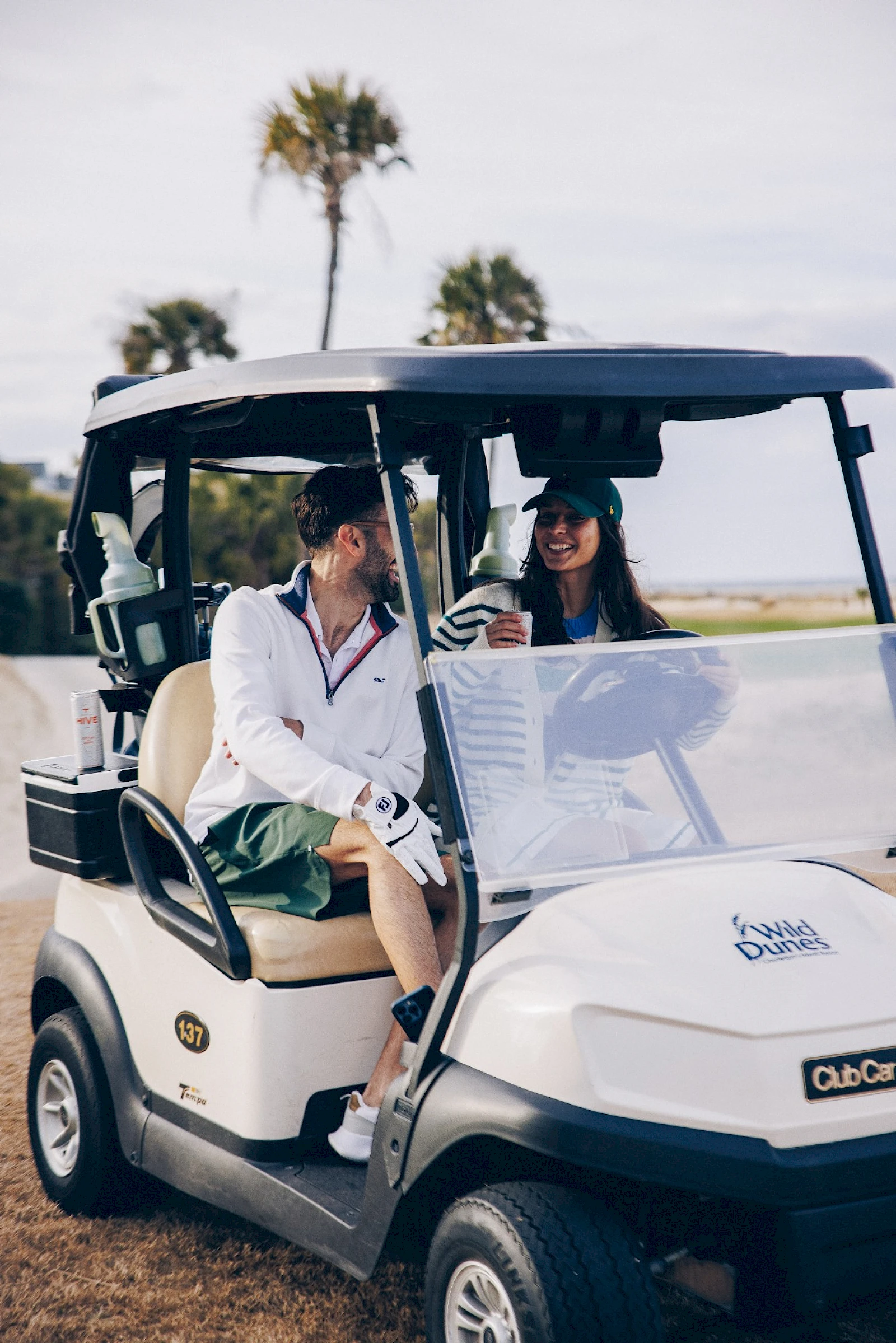 Two people ride in a white golf cart, smiling and chatting on a sunny day with palm trees in the background.