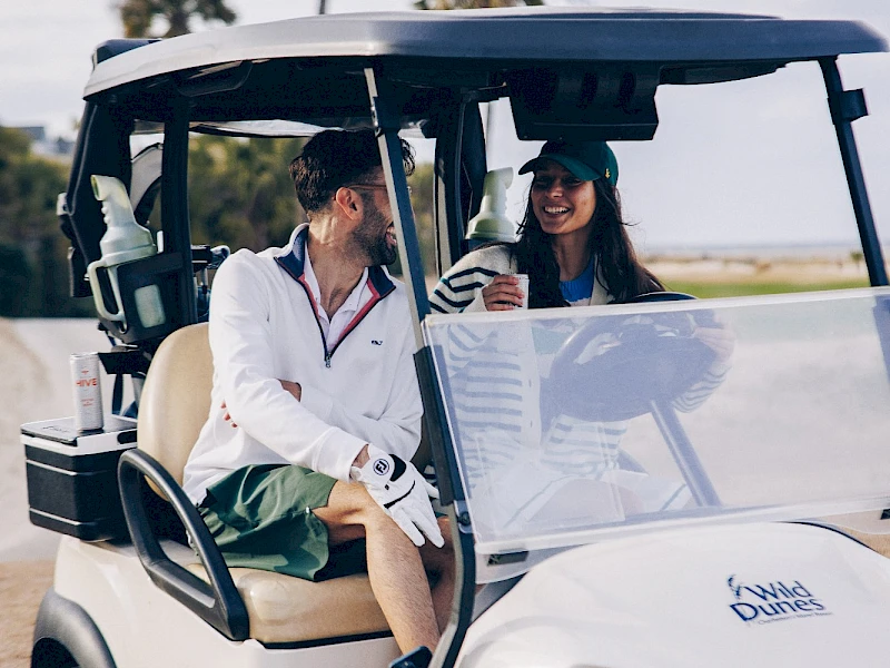 Two people ride in a white golf cart, smiling and chatting on a sunny day with palm trees in the background.