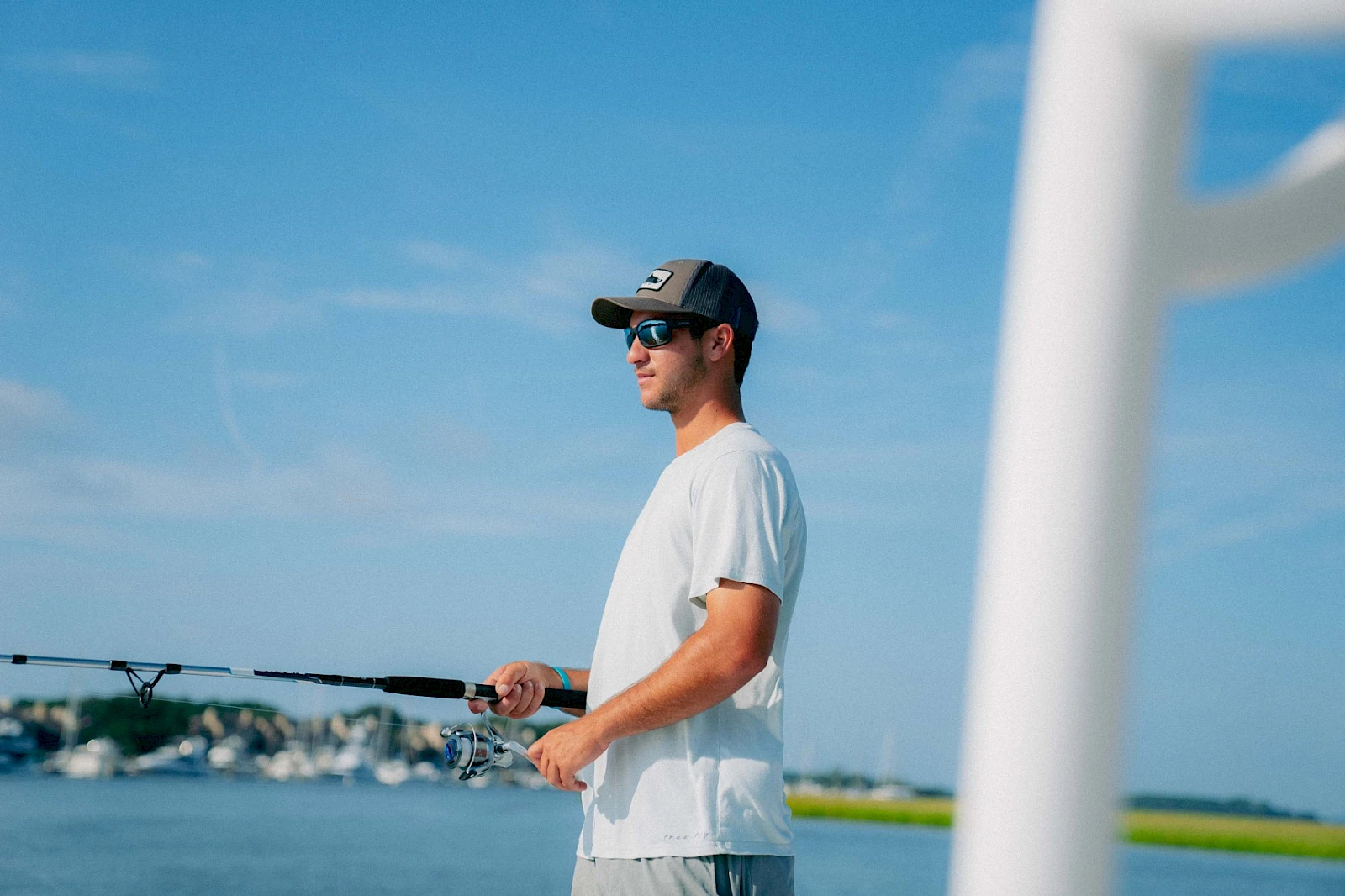 A man wearing a white t-shirt and cap fishing with a rod near a calm blue body of water, clear sky, and a white structure in the foreground.