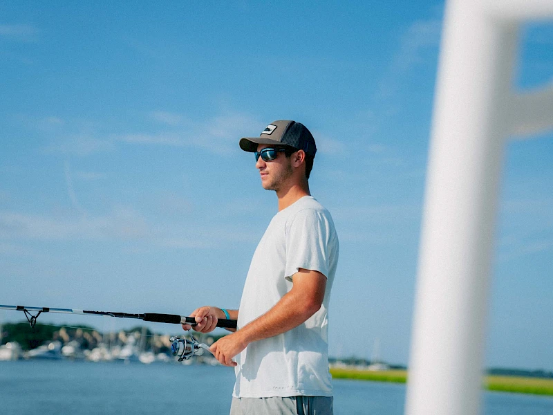 A man wearing a white t-shirt and cap fishing with a rod near a calm blue body of water, clear sky, and a white structure in the foreground.