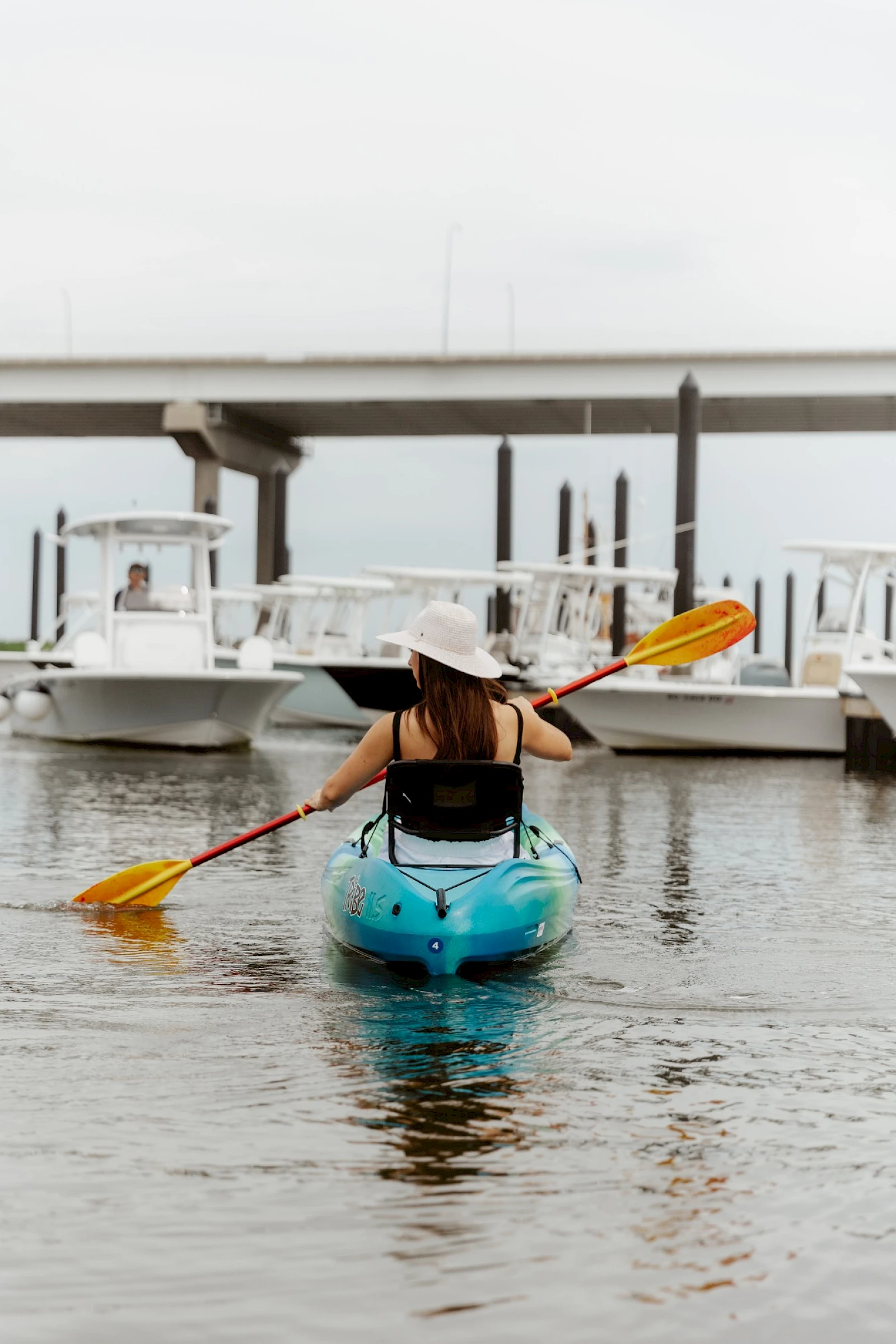 A person paddling a blue kayak toward a marina, wearing a hat and life vest, with boats and a bridge in the background.