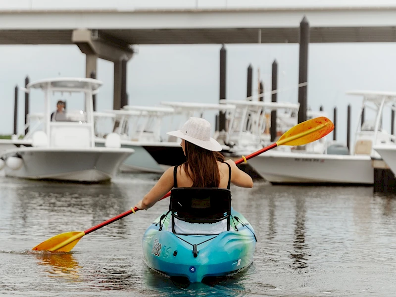 A person paddling a blue kayak toward a marina, wearing a hat and life vest, with boats and a bridge in the background.