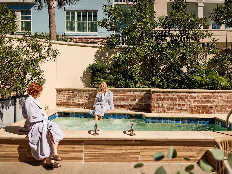 Two people sit by a small pool in robes, relaxing by the spa area in a sunny courtyard.