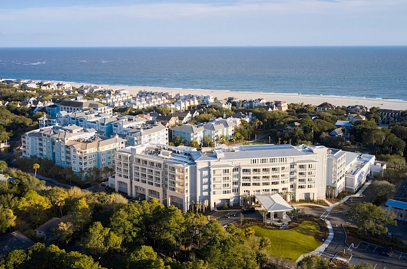 An oceanfront resort town with white multi-story hotels, sandy beach, and lush green trees; blue Atlantic, calm waves, and coastal neighborhood vibes.