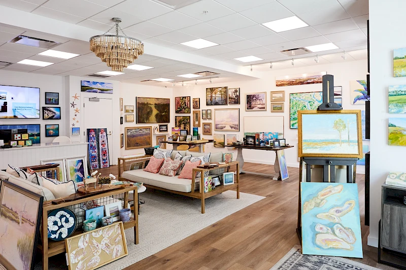 An art gallery or showroom with many paintings on walls, a sofa seating area, display tables, and an easel in the foreground, all warmly lit.