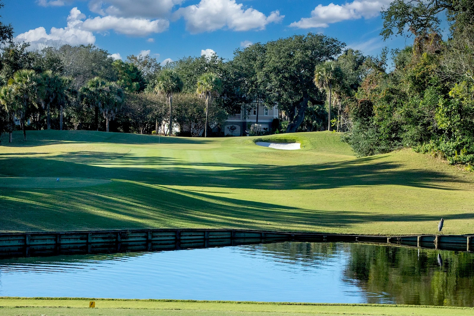 A sunny golf course scene with lush green fairways, a water hazard, trees in the background, and a blue sky with fluffy clouds.