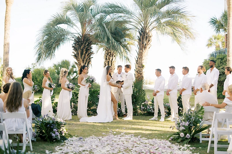 A beachside wedding: bride and groom exchange vows under palm trees, with bridesmaids and groomsmen in white, guests seated and petals on the aisle.