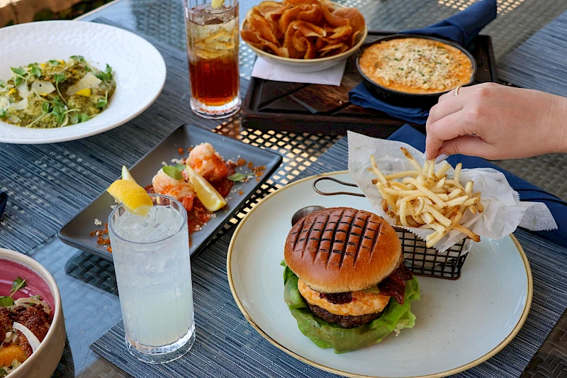 A stylish outdoor spread: a burger with lettuce, fries in a mesh basket, a citrus drink, a small plate of tapas, a salad, and a cheese dip with chips, all on a blue table.