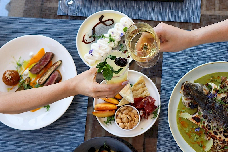 A shared feast: two plates with sausages, veggies, and a small bread roll; a central plate with dips, nuts, and fruit; a glass of white wine, all on a blue table.