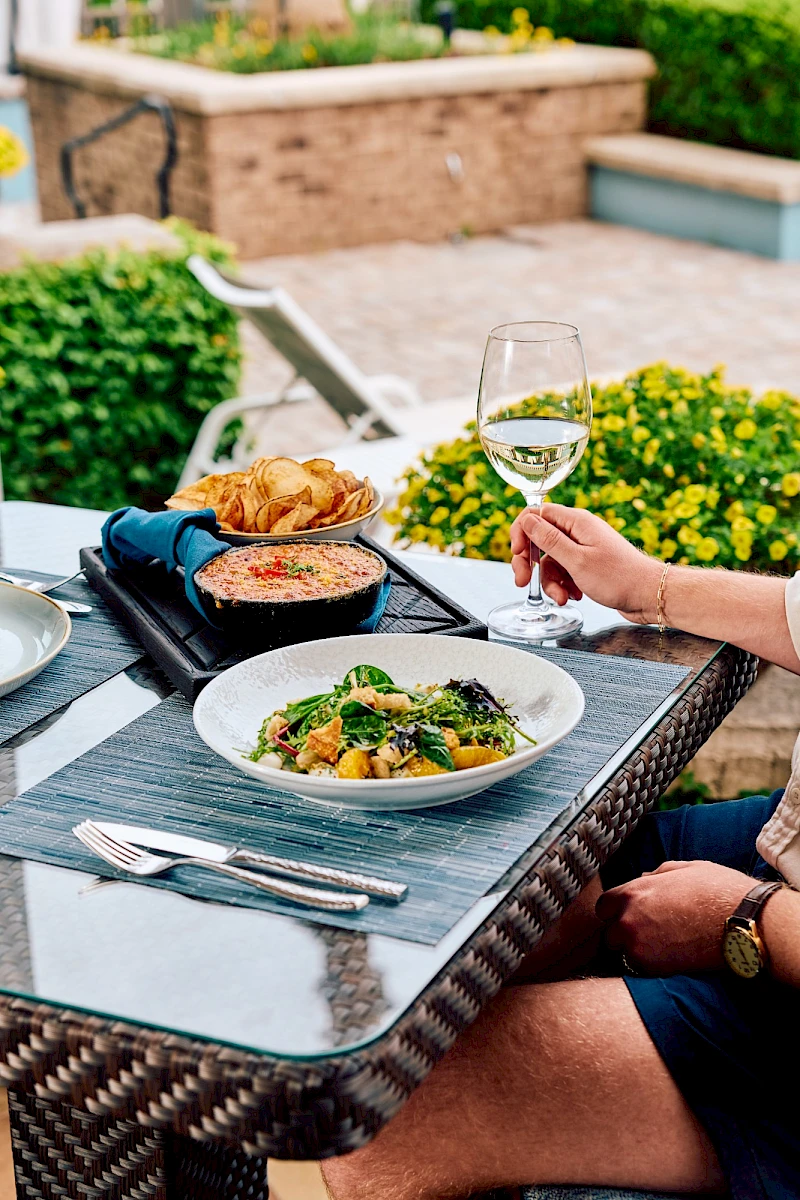 A stylish outdoor lunch: a plate of pasta with greens, a croissant, and a glass of white wine on a glass-top table by a garden—enjoying a sunny meal.