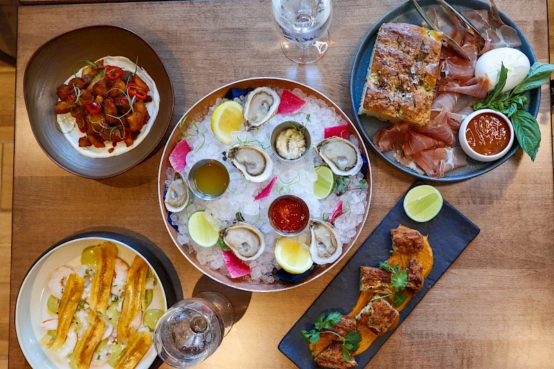 A variety of dishes on a wooden table: a central platter of oysters with lemon wedges and dipping sauces, a plate of calamari with sauces, a pizza slice and greens, a plate of sliders or canapés, and a side dish with crispy breadsticks or crostini, all seen from above.