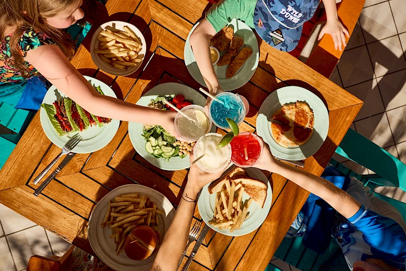 A group of friends around a wooden table sharing a colorful assortment of foods and drinks, seen from above, with hands reaching in to grab snacks.