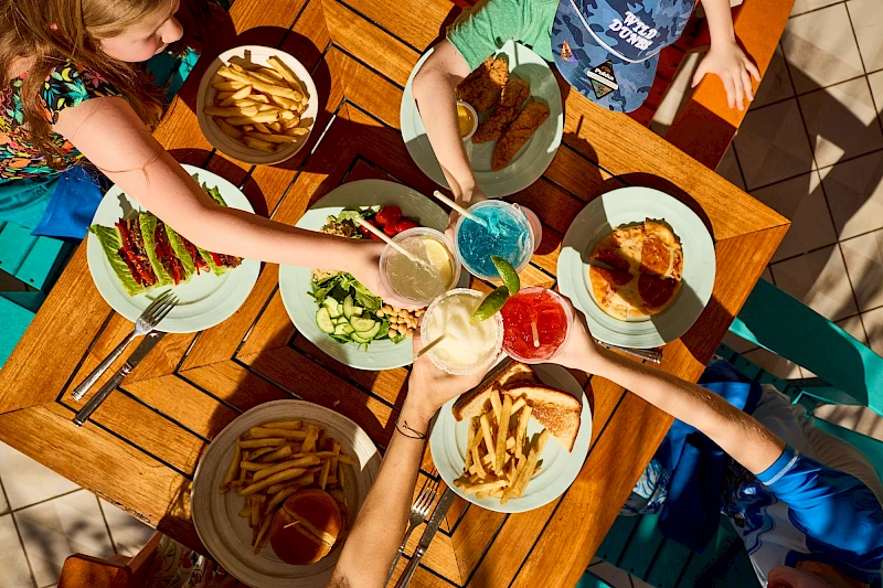 Two women enjoy a sunny outdoor meal at a tropical-style dining area, sharing smiles while eating colorful salads and drinks.