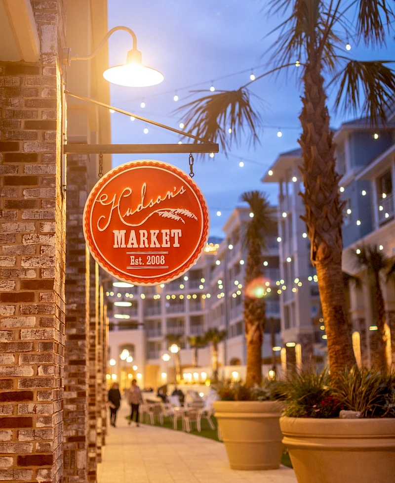A vibrant outdoor market scene at dusk with string lights, palm trees, and a red sign reading “Market” by a brick wall.