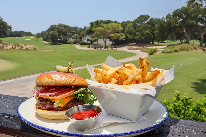 A cheeseburger with lettuce and tomato, a side of french fries in a takeout box, and a small cup of ketchup on a golf-course patio.