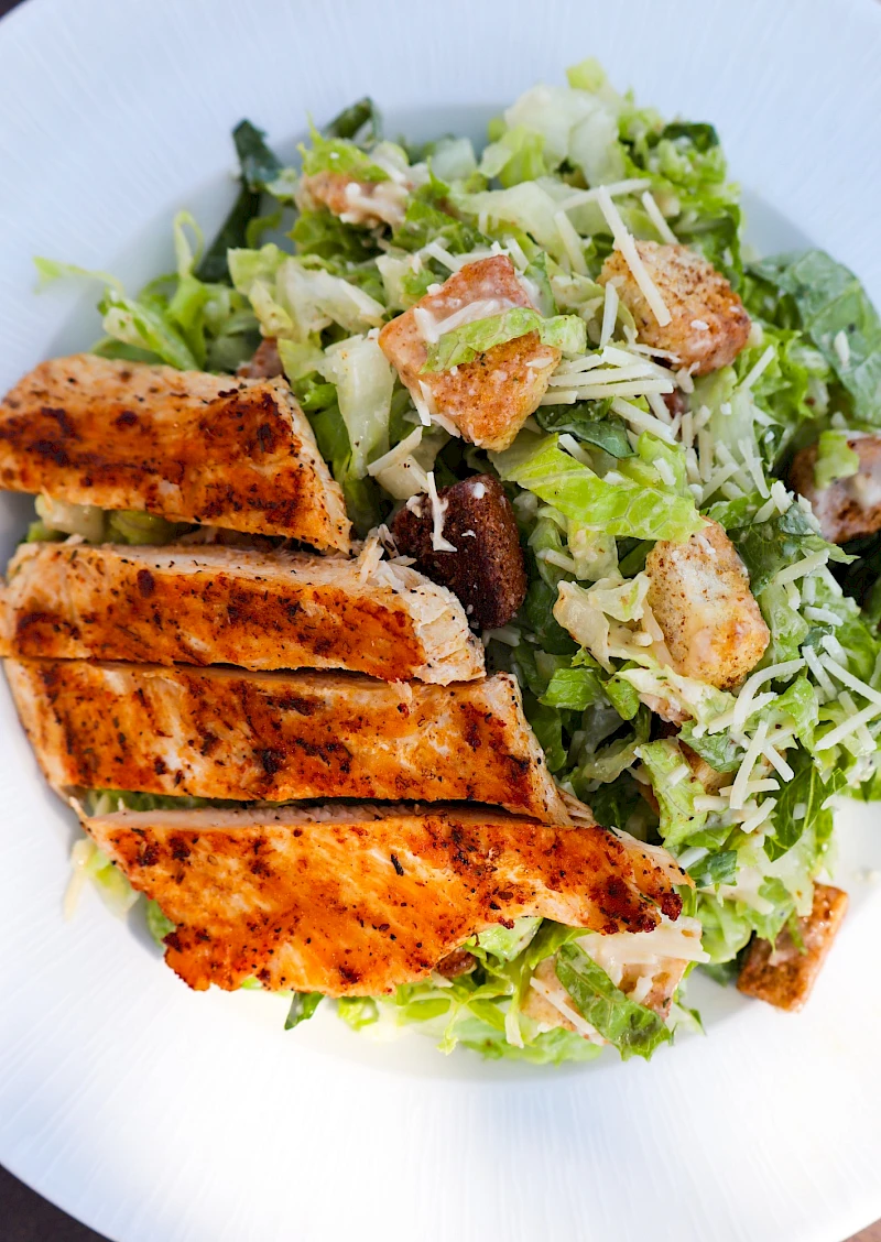 A Caesar-like salad topped with grilled chicken, romaine, croutons, and shaved cheese on a white plate.