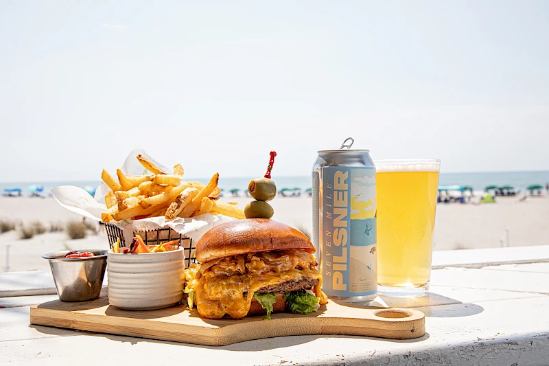 A juicy burger with cheese, crispy fries, a can of Celsius energy drink, and a glass of beer on a wooden board at the beach.