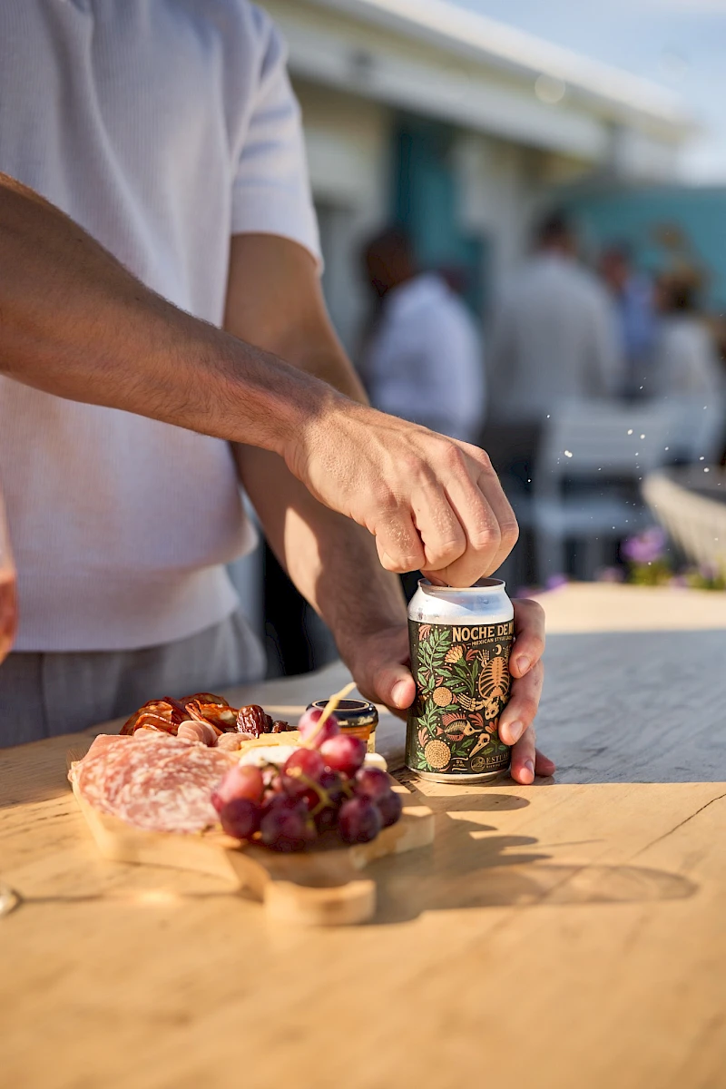 Someone pops a can of beer beside a plate of meat and grapes on a sunny outdoor table.