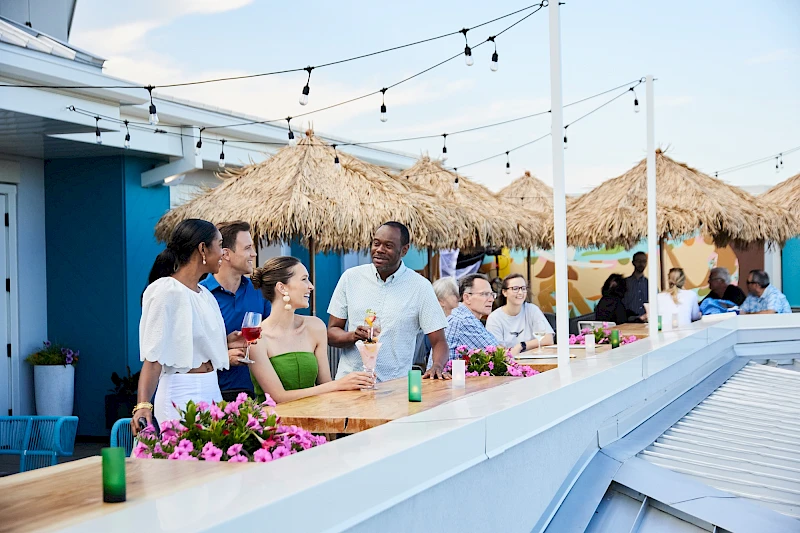 A group of friends enjoy drinks and conversation at an outdoor rooftop bar with string lights and tropical décor, vibrant flowers, and thatched umbrellas.