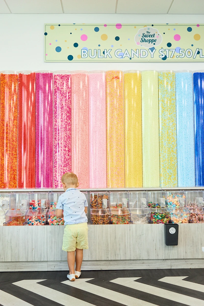 A small child in a light blue shirt and beige shorts stands at a candy counter beneath tall, rainbow-colored candy sticks behind glass.