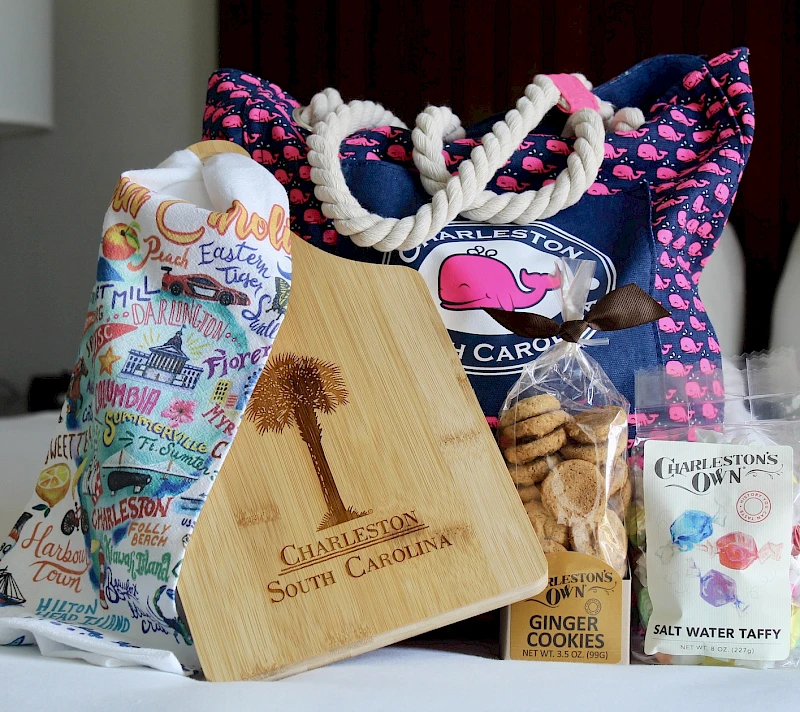A festive gift spread: patterned tote bags, a wooden cutting board engraved “Charleston South Carolina,” cookies, and assorted treats on a table.