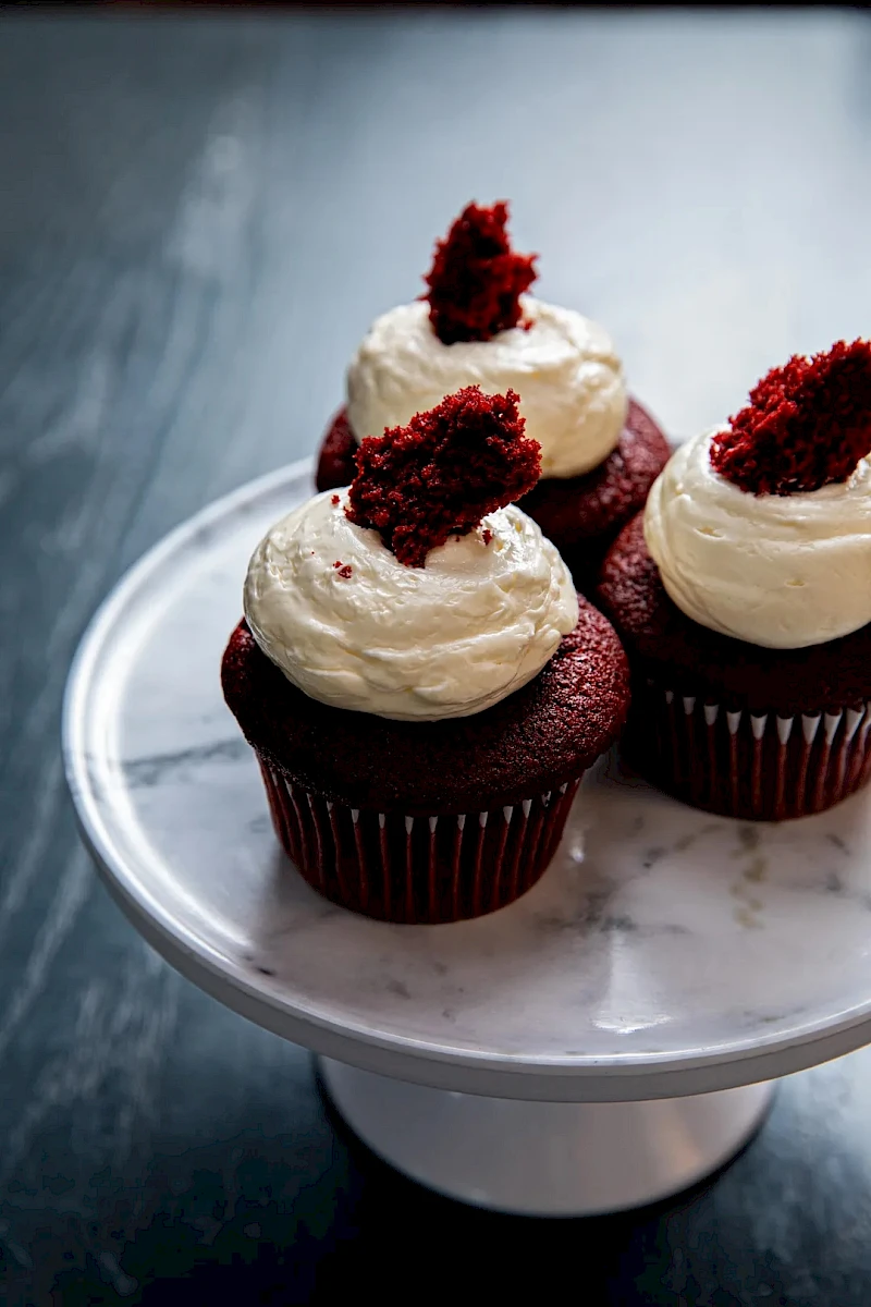 Three chocolate cupcakes topped with swirls of white cream frosting and red cake crumbs on a white cake pedestal.