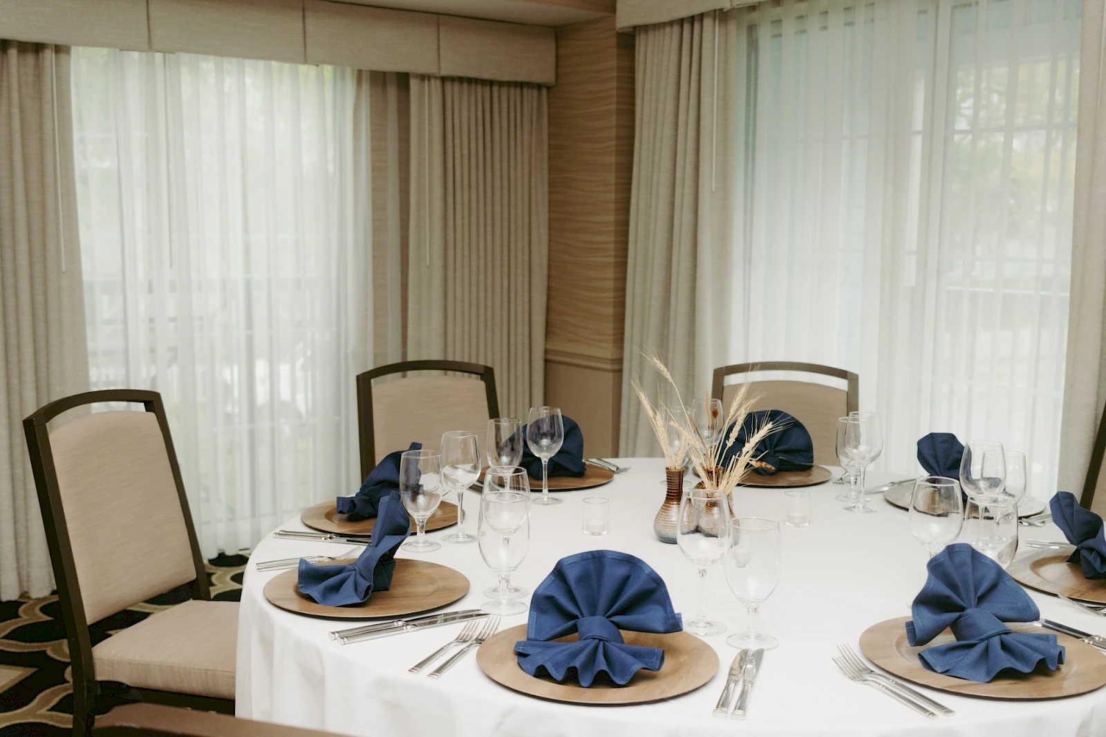 A round table set for dining with white tablecloths, navy napkins folded like flowers, plates, silverware, glasses, and a bright, airy room.
