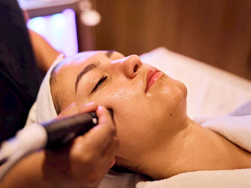 A woman is receiving a facial treatment with a handheld device gliding over her cheek, eyes closed, in a spa-like setting.