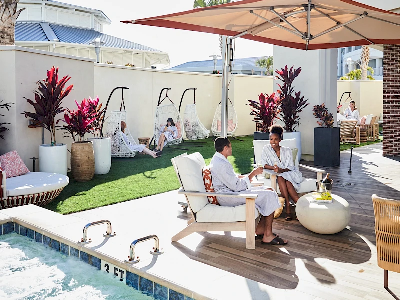A stylish poolside lounge: people relaxing on chairs, white furniture, plants, shade umbrellas, drinks, and a sunlit outdoor patio by the pool.