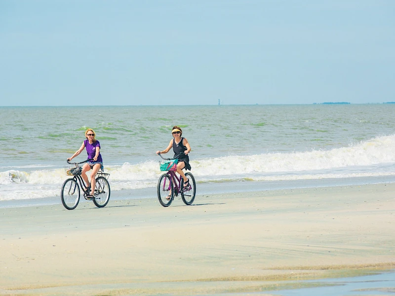 Two people ride bicycles along a sandy beach by the waves, enjoying a sunny day at the coast.