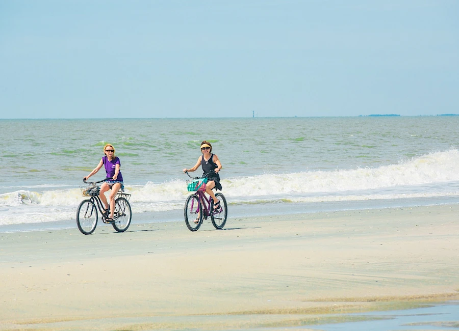 Two people ride bicycles along a sandy beach by the waves, enjoying a sunny day at the coast.