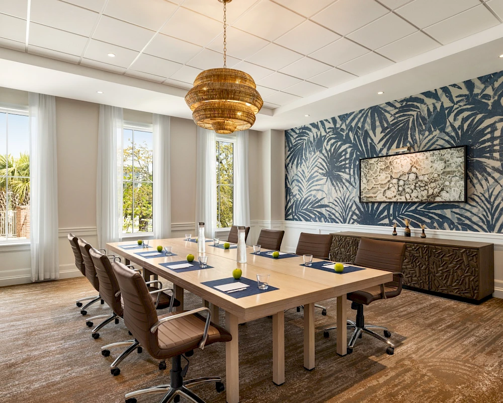 A bright, modern conference room with a long wooden table, eight chairs, tropical leaf wallpaper, large windows, and a woven pendant light hanging above the table.
