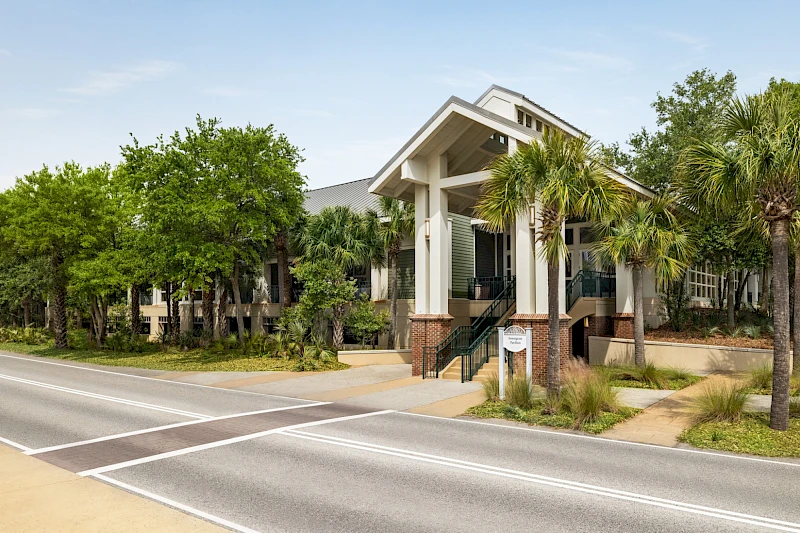 Aiz a modern building with a pitched entrance, palm trees, greenery, and a crosswalk/road in front, reminiscent of a campus or community center.