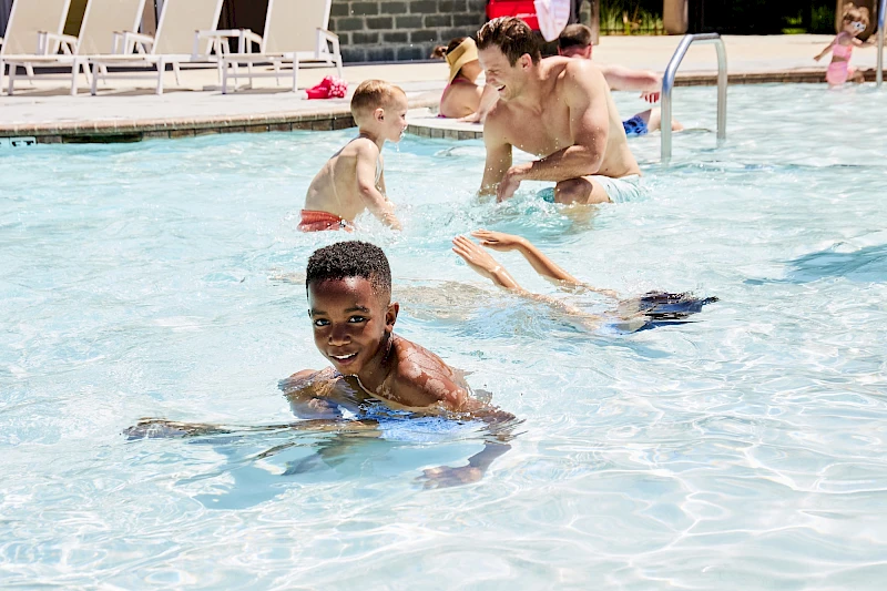 Small kids and adults swim and play in a sunny outdoor pool, splashing and having fun together.