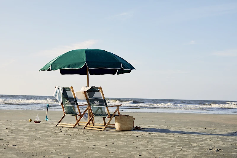 Two beach chairs with a green umbrella sit on the sand, facing the waves, with a small bag nearby and a couple of toys at the edge of the shore.
