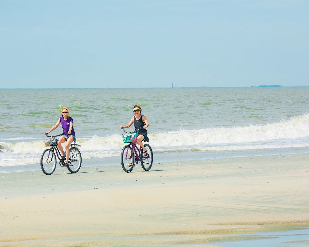Two people ride bicycles along a calm beach shoreline, waves in the background under a clear sky.