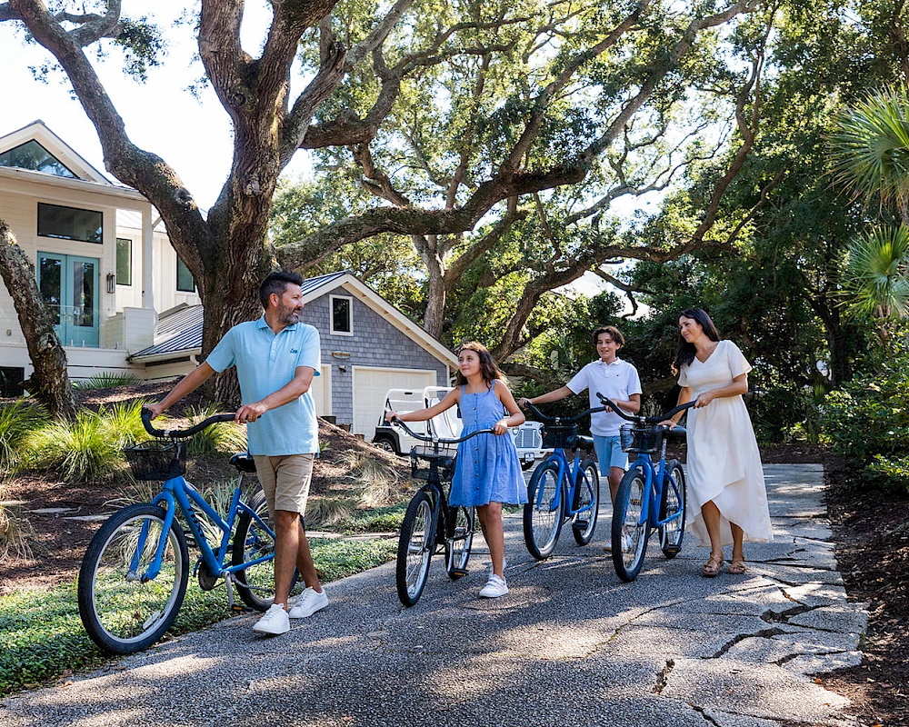 A family of four with bikes stands on a sunny path near a cottage, chatting and ready for a ride among trees and greenery.