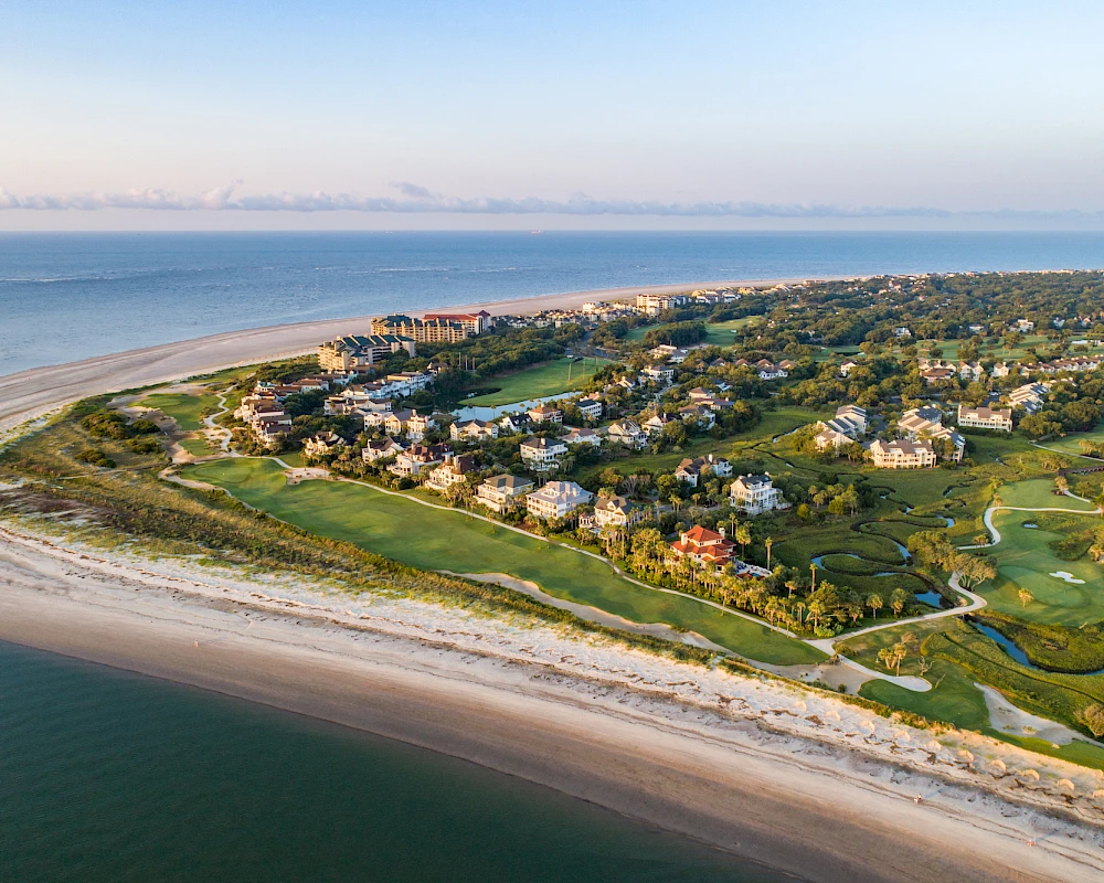 An aerial view of a coastal enclave with sandy beaches, a curved shoreline, residential buildings, and lush green golf-like greens dotted along the coast.