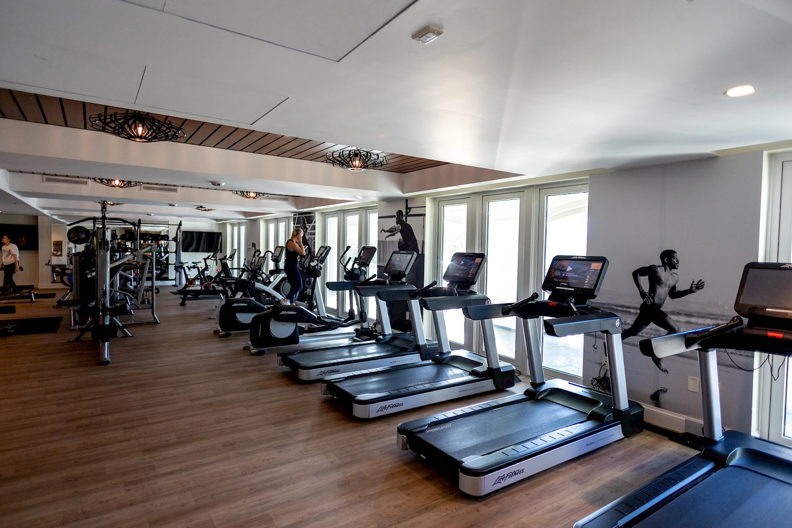 People working out on treadmills in a bright gym with mirrors, weights, and several windows. End with a period.