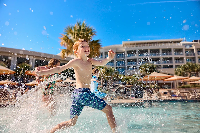 A cheerful young boy runs and splashes through a hotel pool, water spraying around him, with a sunny resort backdrop and palm trees.