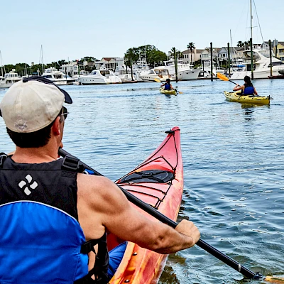 A person in a blue life vest paddles a kayak on calm water near a harbor with sailboats and other kayakers in the distance.