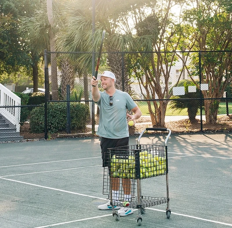 A man in a gray shirt and shorts stands on a tennis court, pushing a shopping cart filled with tennis balls, waving or gesturing.