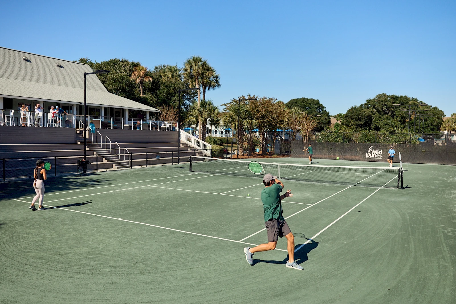 Two men play tennis on a green outdoor court with a cheering group in a clubhouse area and palm trees in sunny weather, ending with a period.